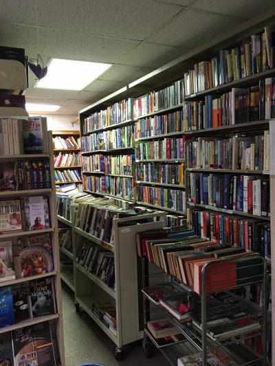 Dimly lit cozy bookstore aisle with shelves and carts full of colorful books. The setting feels inviting and intimate, perfect for browsing.