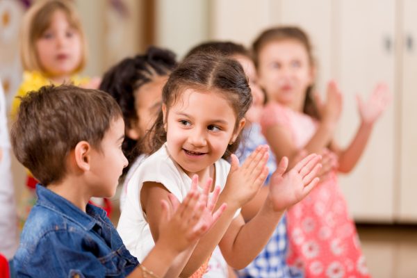 A group of young children clapping, with a smiling girl in the foreground looking at a boy. The scene is lively and joyful, set indoors.