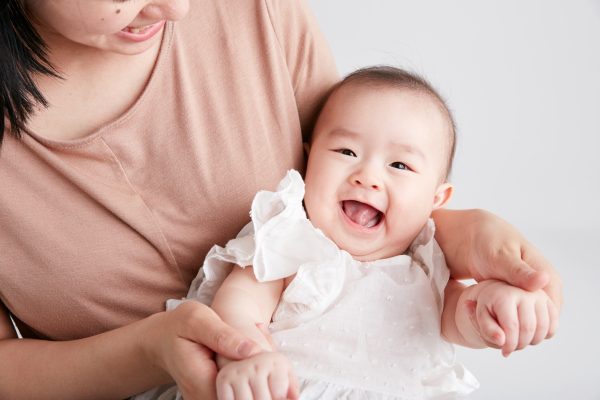 A smiling baby in a white outfit is being held by an adult in a peach top. The scene conveys warmth and joy, set against a soft gray background.