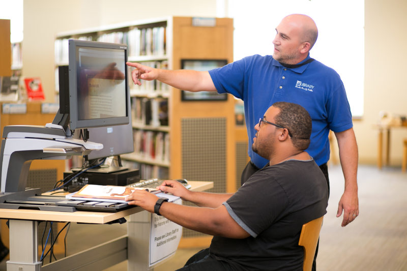 A library staff member in a blue shirt helps a man using a computer with a magnified screen, pointing to assist, creating a focused and helpful atmosphere.