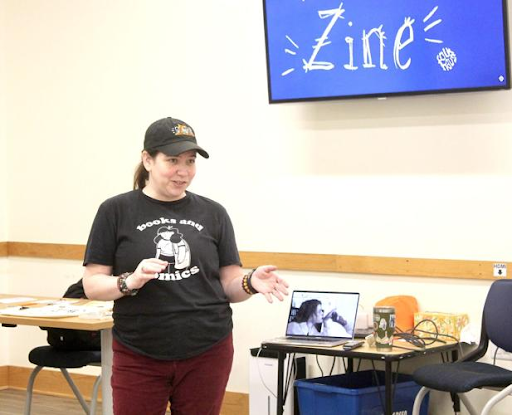 A woman in a black t-shirt and baseball cap speaks in front of a screen displaying the word Zine, with a laptop and desk visible in the room.