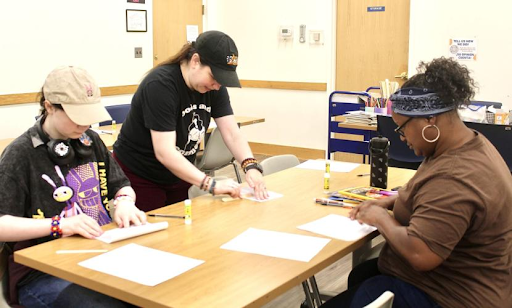 Three people sit at a large wooden table in a room, working on craft projects with paper, glue sticks, and markers spread across the surface.