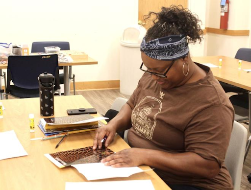 A woman wearing a brown shirt and a patterned headband sits at a table, looking down while gluing a magazine cut-out down in a bright classroom.